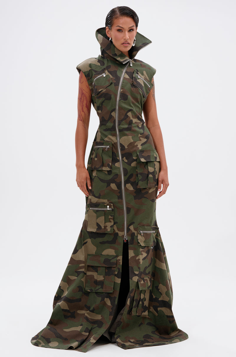 A person stands confidently in the NOW YOU SEE ME CAMO PRINT MAXI DRESS, featuring a high collar, zipper front, and multiple cargo pockets, set against a plain white background.