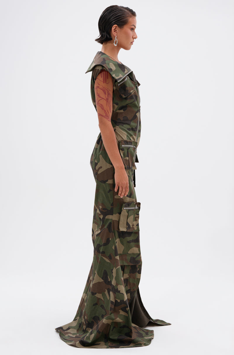 A person with short, slicked-back hair and a tattooed arm stands in profile wearing the NOW YOU SEE ME CAMO PRINT MAXI DRESS featuring a large collar, cargo pockets, and a subtle train.