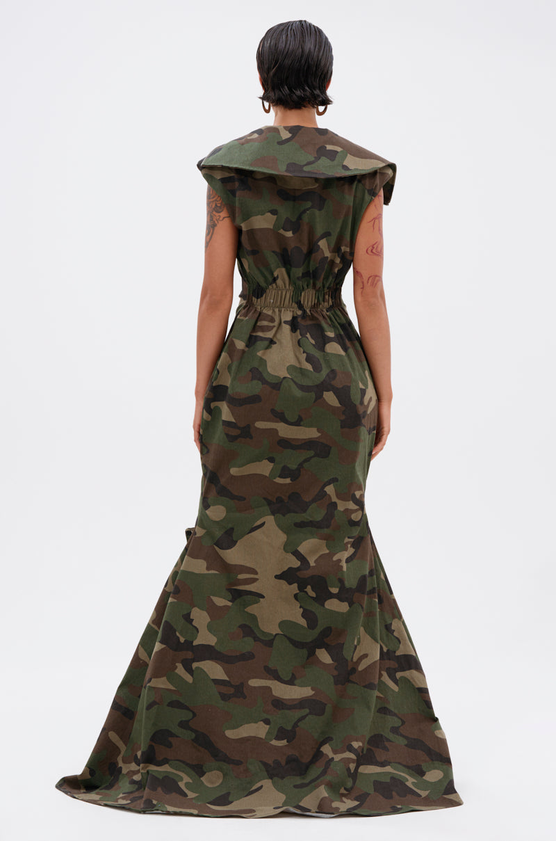 A person with short dark hair faces away while wearing the NOW YOU SEE ME CAMO PRINT MAXI DRESS, featuring a fitted waist, flared hem, and cargo pockets, set against a plain white background.