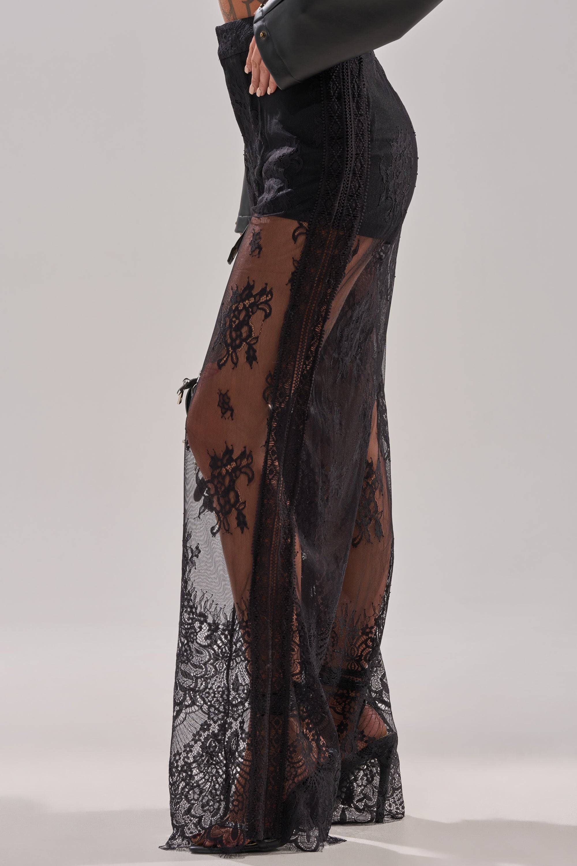 A person is wearing the MISS ME YET LACE TROUSER—high-waisted, sheer black lace pants with intricate floral patterns and solid black panels—paired with a black top. The focus is on the elegant trousers from waist to floor.
