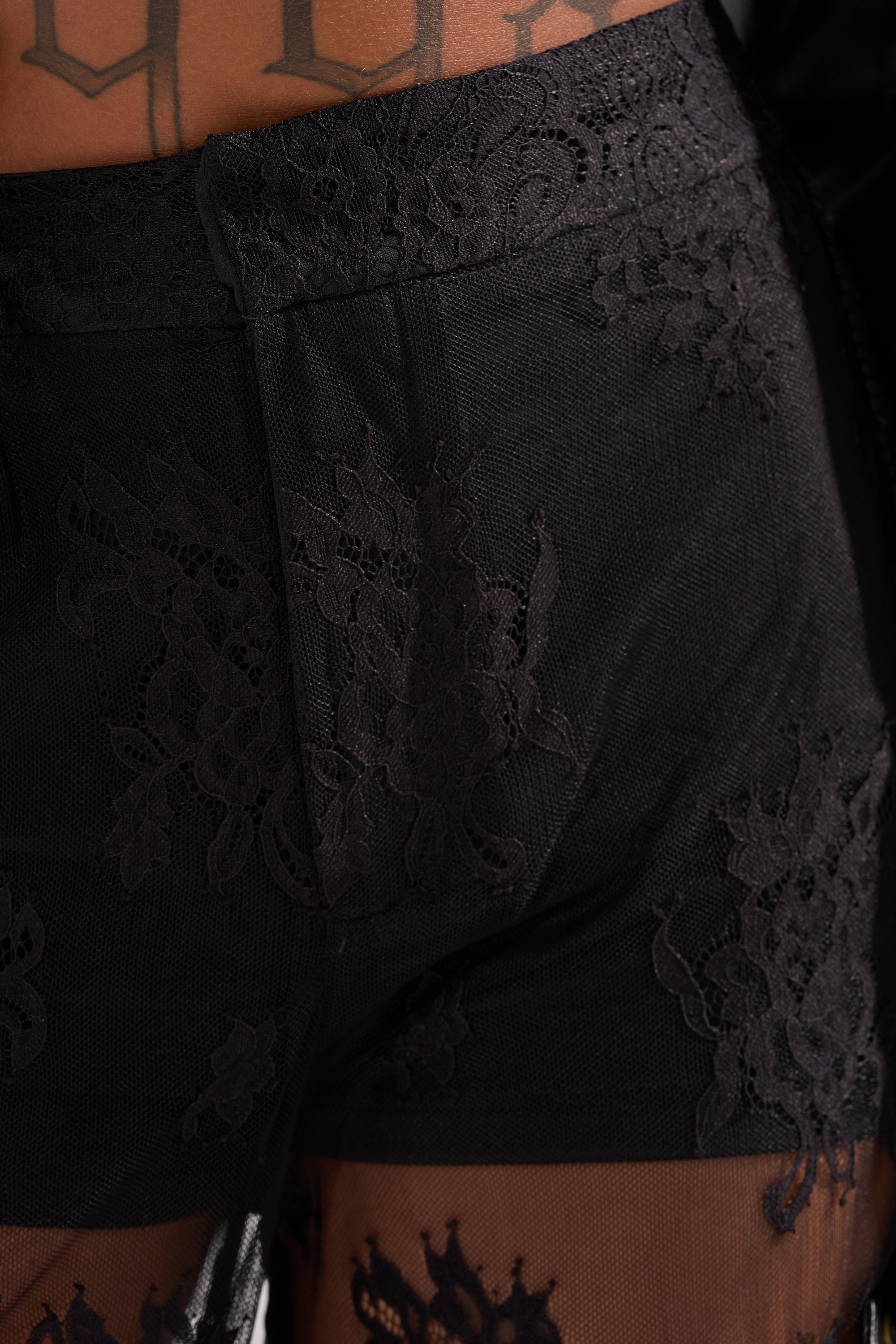 Close-up of a person wearing the MISS ME YET LACE TROUSER—black, see-through shorts with a sheer floral lace pattern. The delicate lace reveals skin beneath, and part of a bold black-lettered tattoo is visible on their lower torso.
