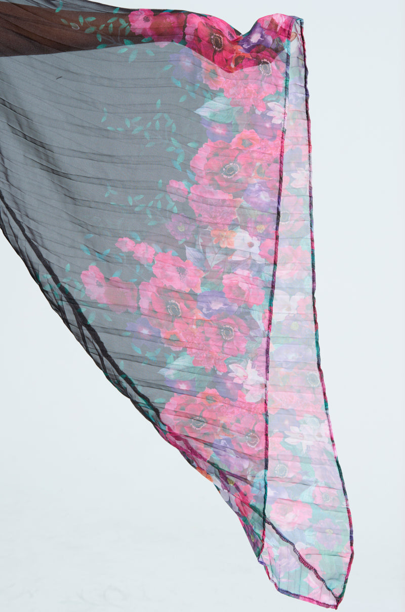 THE JULIE BLOUSE features a sheer black fabric with a tie-front closure and a vibrant floral pattern in pink, purple, and green. Its flowing material highlights both its translucent quality and intricate flower design.