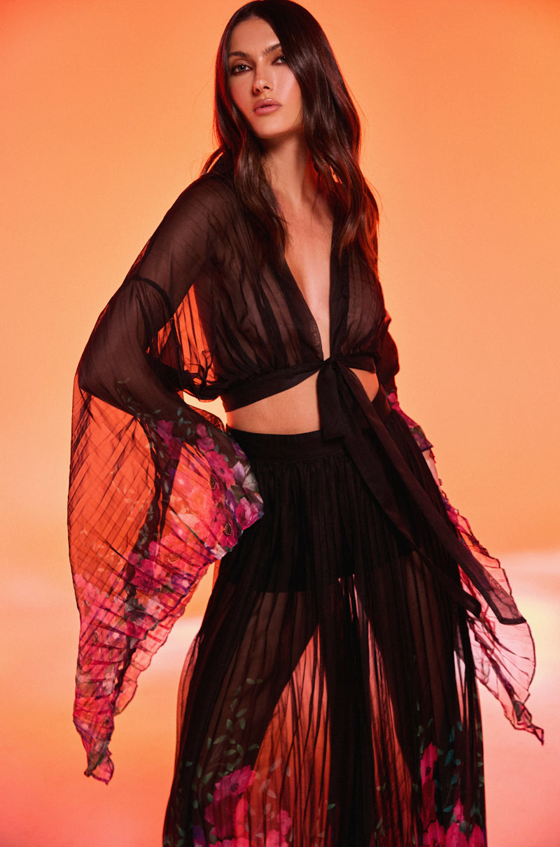 A woman poses against an orange background wearing THE JULIE BLOUSE, a sheer mesh top with floral-patterned sleeves and hem, a deep V-neck, waist ties, and wide flowing sleeves for a dramatic look.