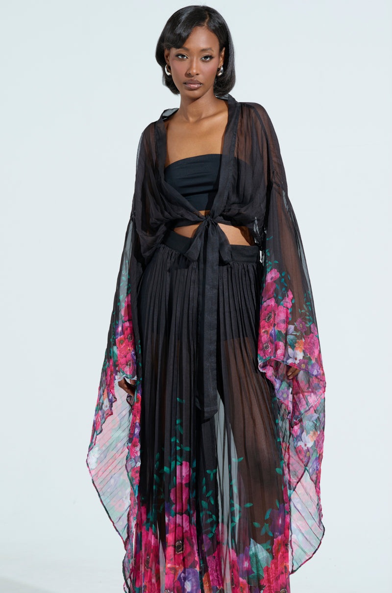 A woman wears THE JULIE BLOUSE, a black sheer mesh outfit with vibrant pink and purple floral designs. The tie-front cropped blouse enhances her style as she stands against a light gray background.