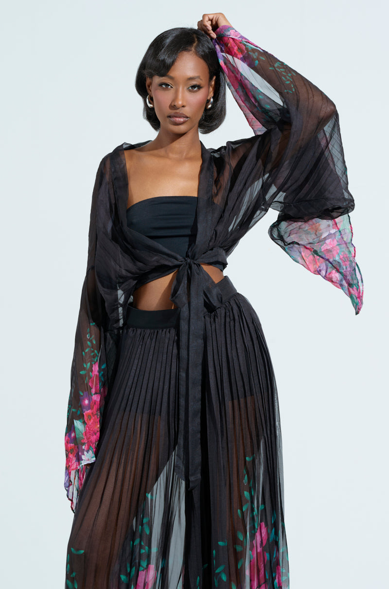 A woman confidently poses in THE JULIE BLOUSE, featuring a sheer black, floral pattern. The flowy top and skirt reveal a black tube top and shorts underneath, with one arm raised to showcase the pleated fabric.