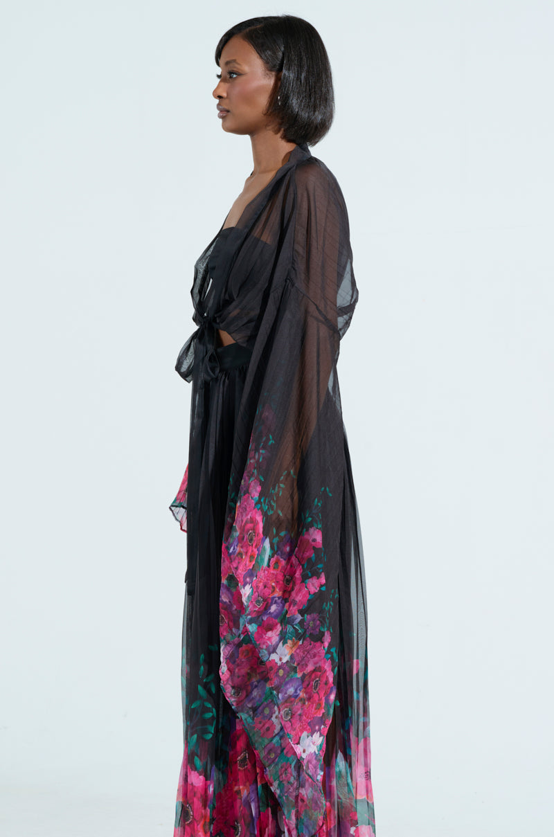 In profile, a woman wears THE JULIE BLOUSE, featuring flowing black fabric with pink and red floral patterns at the bottom, reminiscent of a tie-front cropped design, set against a plain light background.