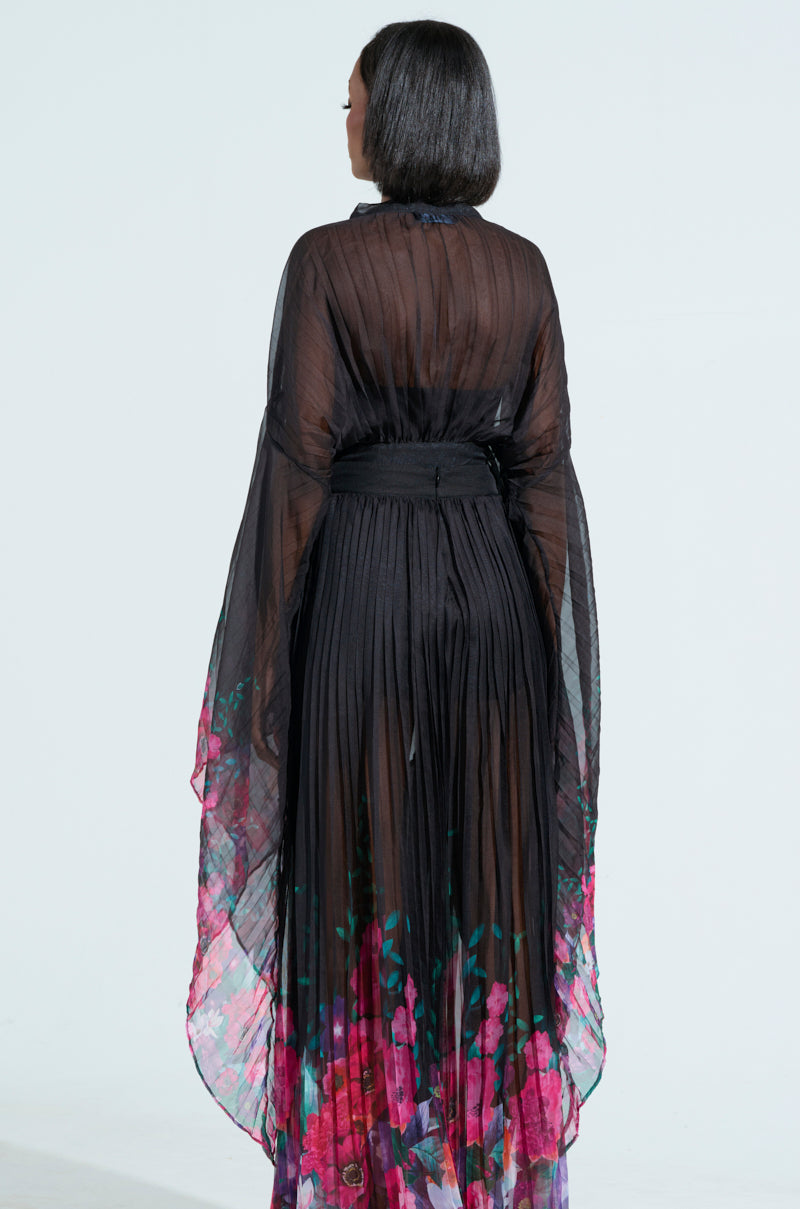 A person with straight black hair is seen from behind wearing THE JULIE BLOUSE—an elegant, long black gown with wide sleeves and a sheer pleated design that features a vibrant floral pattern at the bottom, creating a flowing effect.