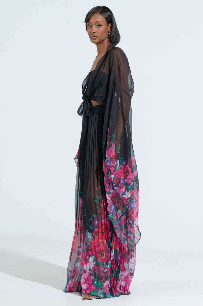 A model poses against a light background in THE JULIE BLOUSE, a sheer, black floral piece adorned with vibrant pink and purple flowers. The blouse features long, flowing sleeves and a tie-front design, creating an elegant, layered appearance reminiscent of a gown.