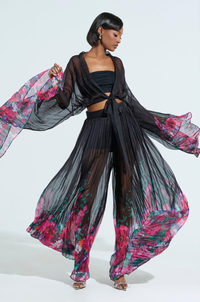 A person elegantly wears THE JULIE BLOUSE, featuring sheer mesh with vibrant pink and red floral patterns on the sleeves and pants. The garment ties at the front, revealing a black top and shorts beneath, as they gracefully hold the flowing fabric aloft.