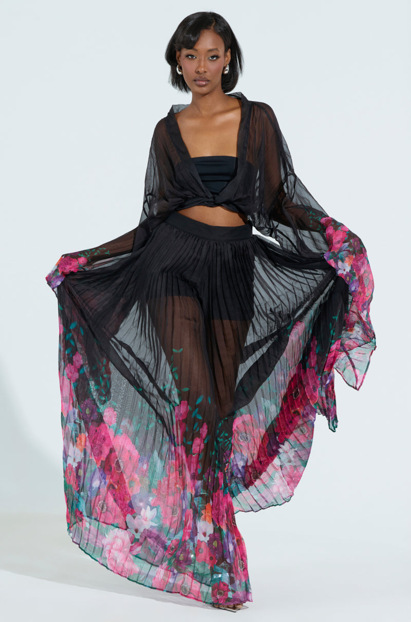 Wearing THE JULIE BLOUSE, a woman pairs it with high-waisted flowy pants, both featuring vibrant pink and purple floral patterns at the hem. She poses gracefully, spreading the fabric for an elegant and dynamic look.