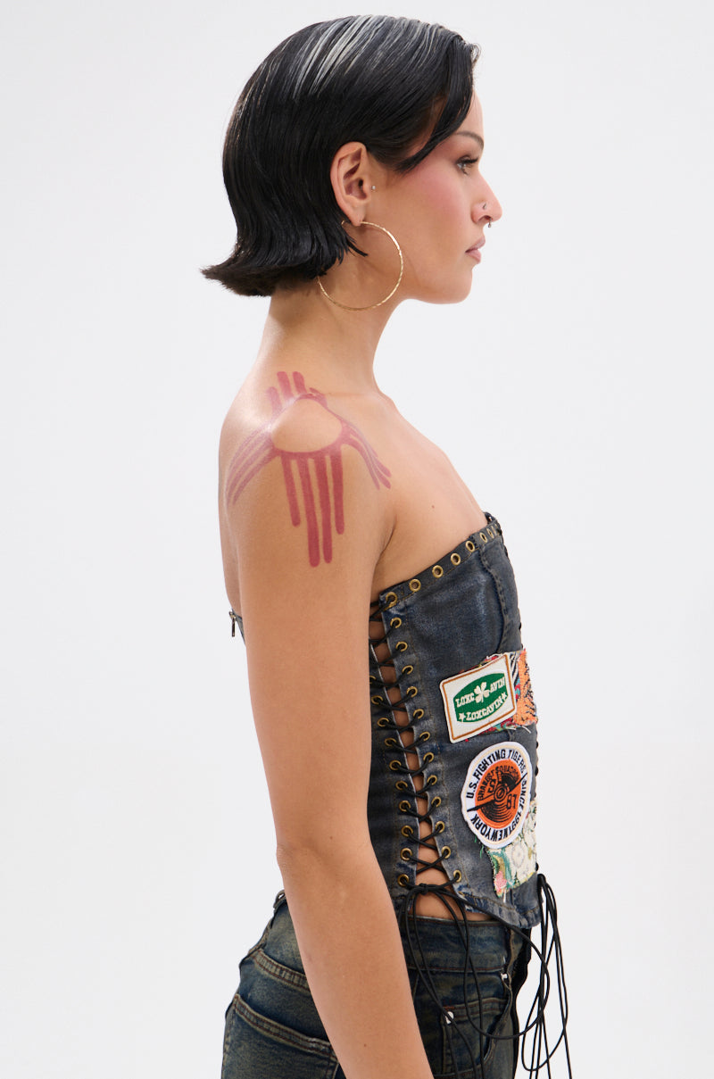 A person with short dark hair and hoop earrings stands in profile, wearing the ARTS AND CRAFTS PATCH WORK DENIM CORSET with embroidered patches and pink handprint-like makeup on their shoulder and upper arm.