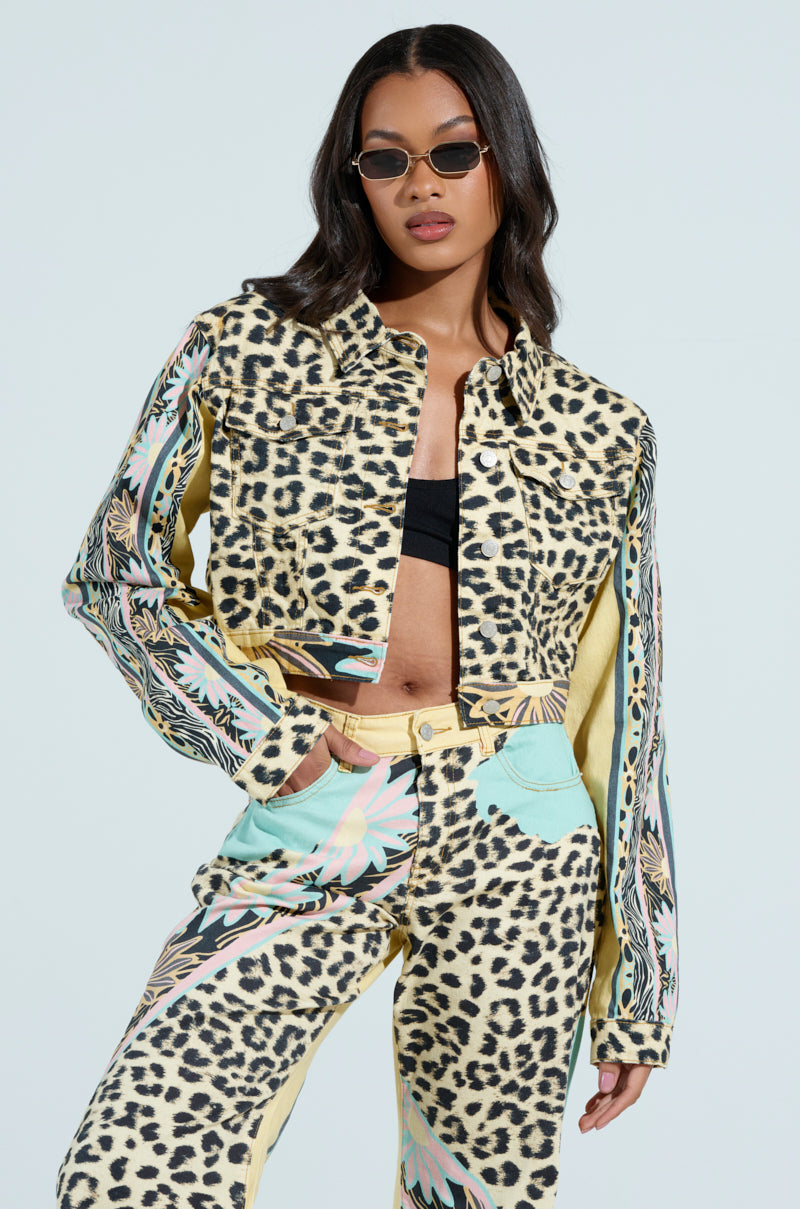 A woman confidently poses in small sunglasses and a leopard-print outfit, showcasing a cropped silhouette with the TRUST YOUR INSTINCTS DENIM JACKET, featuring chic colorful patterns on the sleeves and pockets against a plain background.