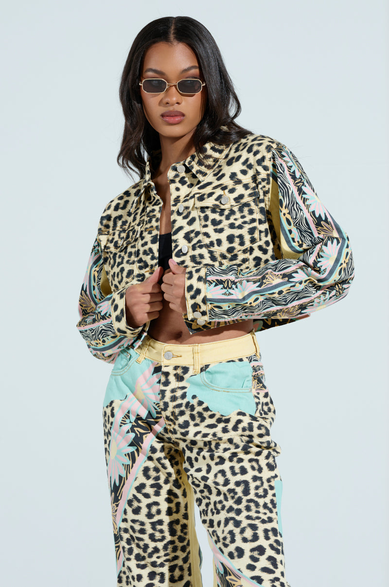 A person confidently poses in sunglasses and a colorful outfit featuring a bold mix of leopard prints and abstract patterns, complemented by the chic TRUST YOUR INSTINCTS DENIM JACKET with pastel accents, set against a plain background.