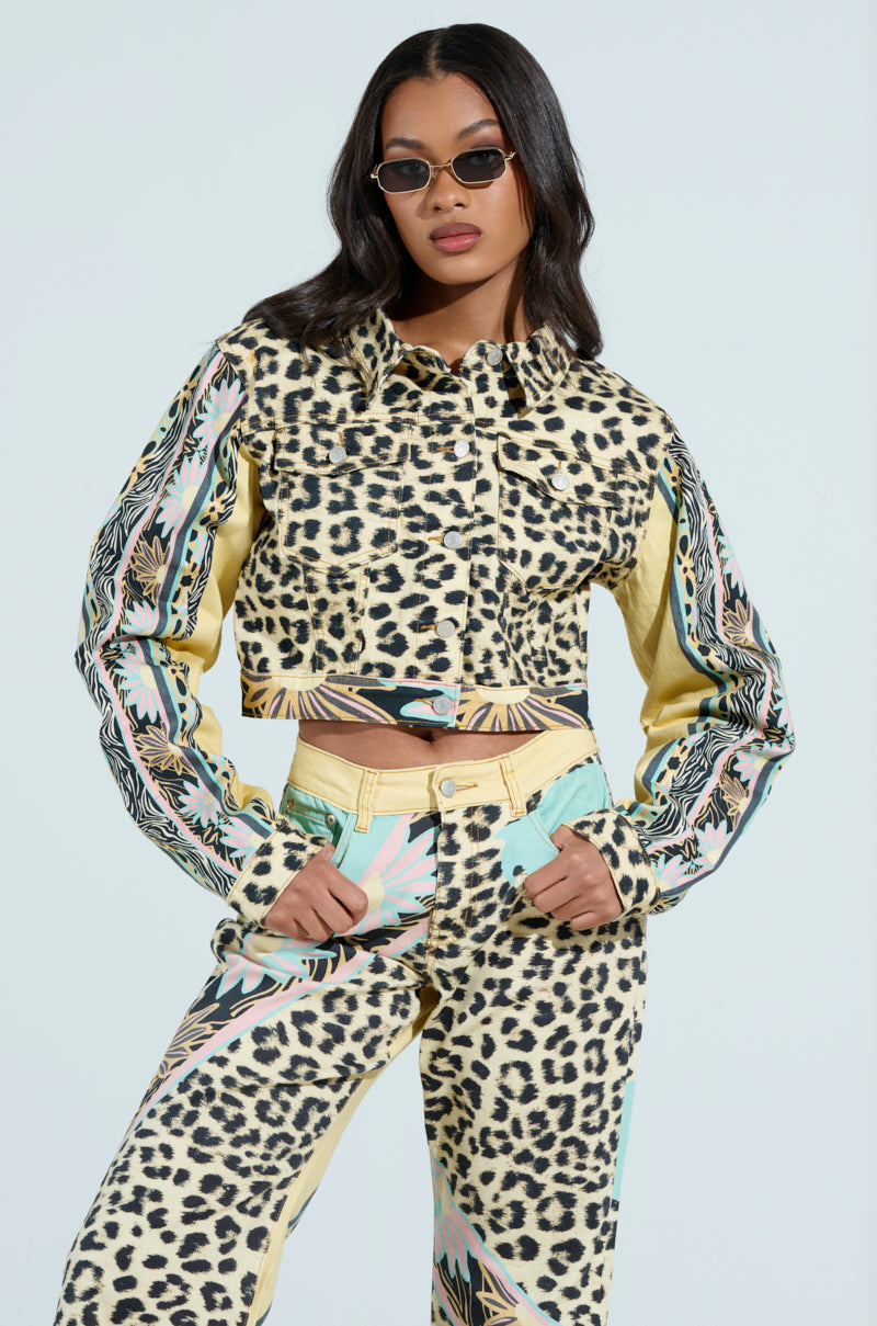 A woman confidently stands with hands in her pockets, wearing the TRUST YOUR INSTINCTS DENIM JACKET and matching leopard-print pants. Her long dark hair pairs perfectly with her sunglasses as she poses against a light blue backdrop, embracing the chic allure of her cropped silhouette.