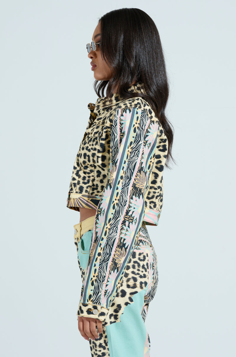 A woman in a TRUST YOUR INSTINCTS DENIM JACKET and vibrant leopard-print outfit with stripes displays a cropped silhouette. Her long dark hair and sunglasses create a bold style against the plain background, perfectly complemented by the matching pants.