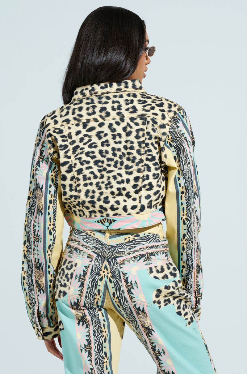 A person with long dark hair models a colorful outfit featuring the cropped TRUST YOUR INSTINCTS denim jacket in leopard print. The pastel-hued pants add intricate designs. Wearing sunglasses, they strike a pose against a light background, exuding effortless style.