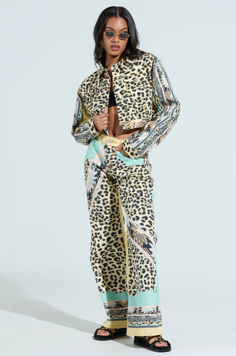 Wearing sunglasses and a leopard-print co-ord with a tropical pattern, a woman stands confidently with one hand in her pocket, draped in the TRUST YOUR INSTINCTS DENIM JACKET over her shoulders. She pairs this look with black sandals against a light blue background.