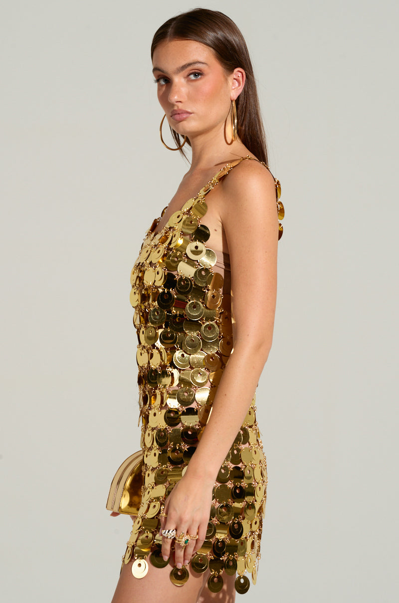 A woman stands in a glamorous HEART OF GOLD MINI DRESS, adorned with oversized sequins, paired perfectly with large hoop earrings. The sleeveless mini-dress matches her small metallic gold chain clutch. Her expression is neutral, and her long, straight hair contrasts the plain light-colored background.