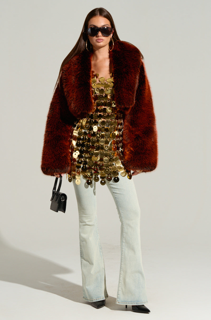 A person dressed in a glamorous HEART OF GOLD MINI DRESS and a fluffy brown jacket, paired with light blue flared jeans. They have long hair, sunglasses, hoop earrings, and are holding a small black handbag.