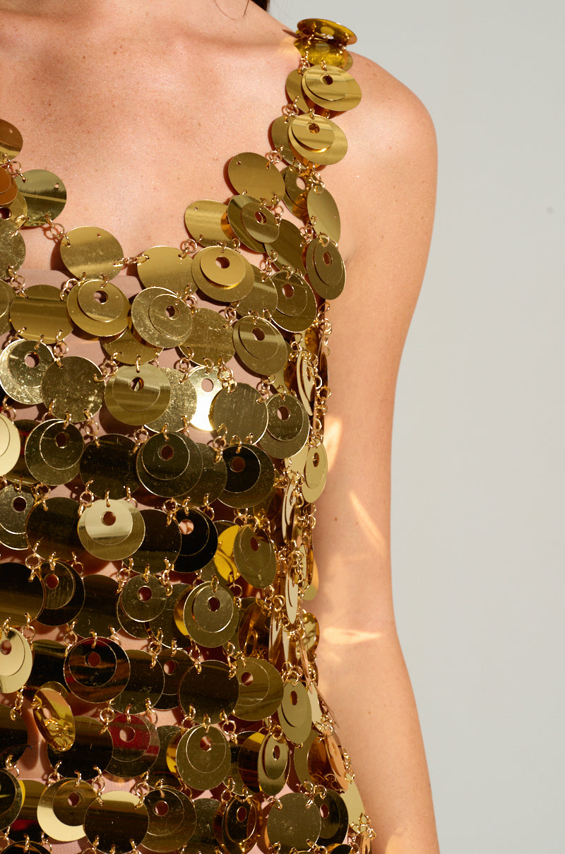 A person wears the HEART OF GOLD MINI DRESS against a neutral background. The glamorous, overlapping gold discs reflect light, adding texture and shimmer to the ensemble. The image focuses on the upper body and arm, highlighting the dress's reflective allure.