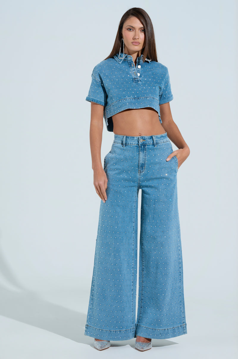 Against a light blue backdrop, a woman exudes style in a cropped denim shirt with rhinestones, paired with IT'S GIVING DENIM PANT wide-leg jeans and silver shoes. Her right hand in the pocket emphasizes her chic ensemble and long, straight hair.