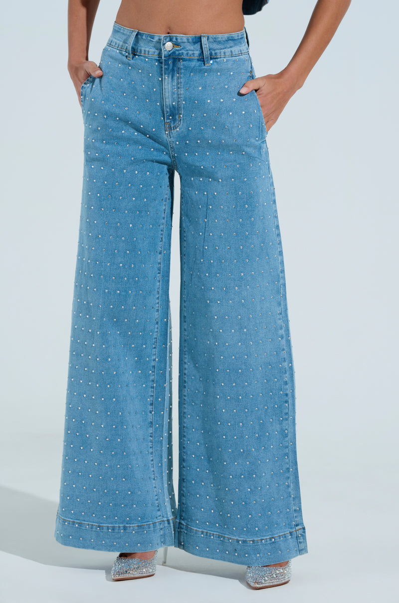 Someone is wearing the IT'S GIVING DENIM PANT featuring high-waisted, wide-leg denim with a dotted pattern and rhinestone details. Their hands are in pockets and they pair the jeans with sparkly silver shoes, set against a light blue background.