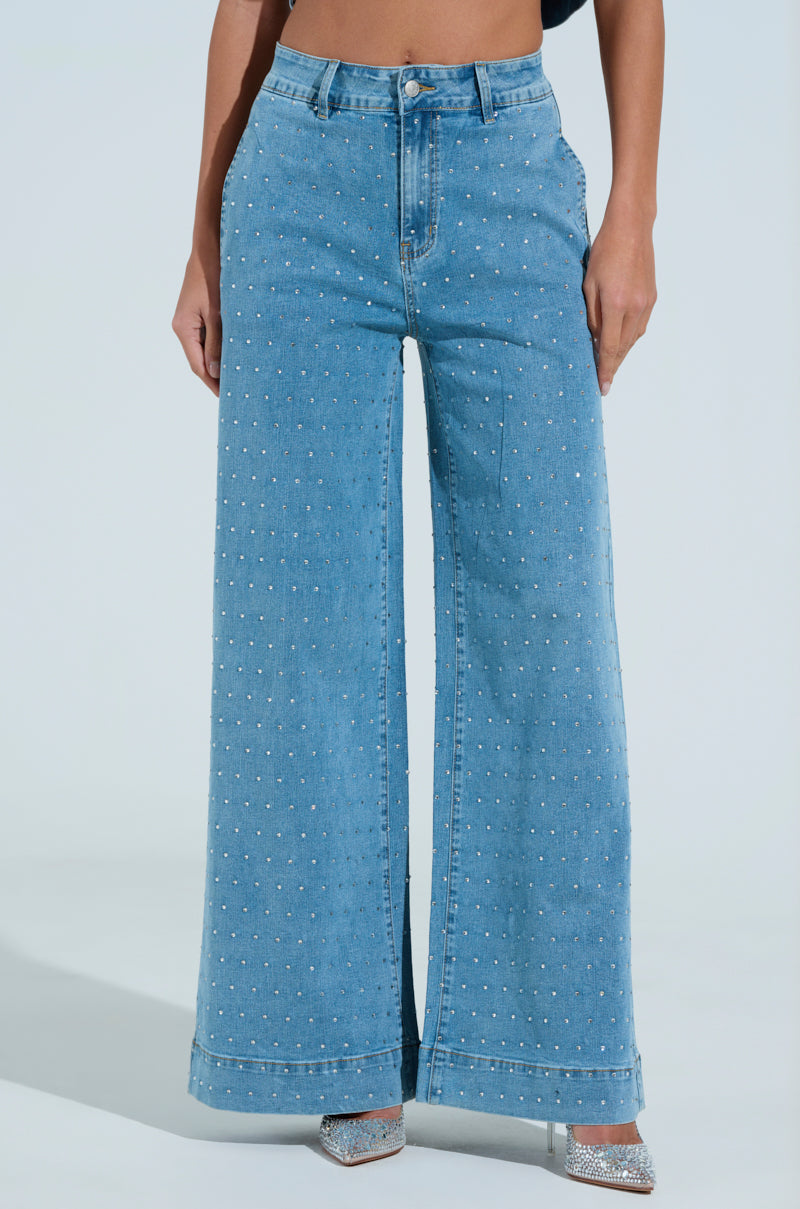 A person wears the "IT'S GIVING" denim pant with a high-waisted, wide-leg design and small white polka dots, paired with sparkly heels adorned with rhinestone embellishments, set against a light blue background.