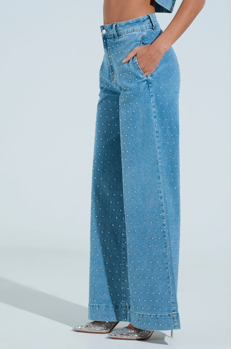 A person is wearing the IT'S GIVING DENIM PANT—a pair of high-waisted, wide-leg jeans with decorative pearls. They're striking a pose with one hand in their pocket, and the ensemble is completed with rhinestone-embellished, sparkly pointed-toe heels against a simple, light background.