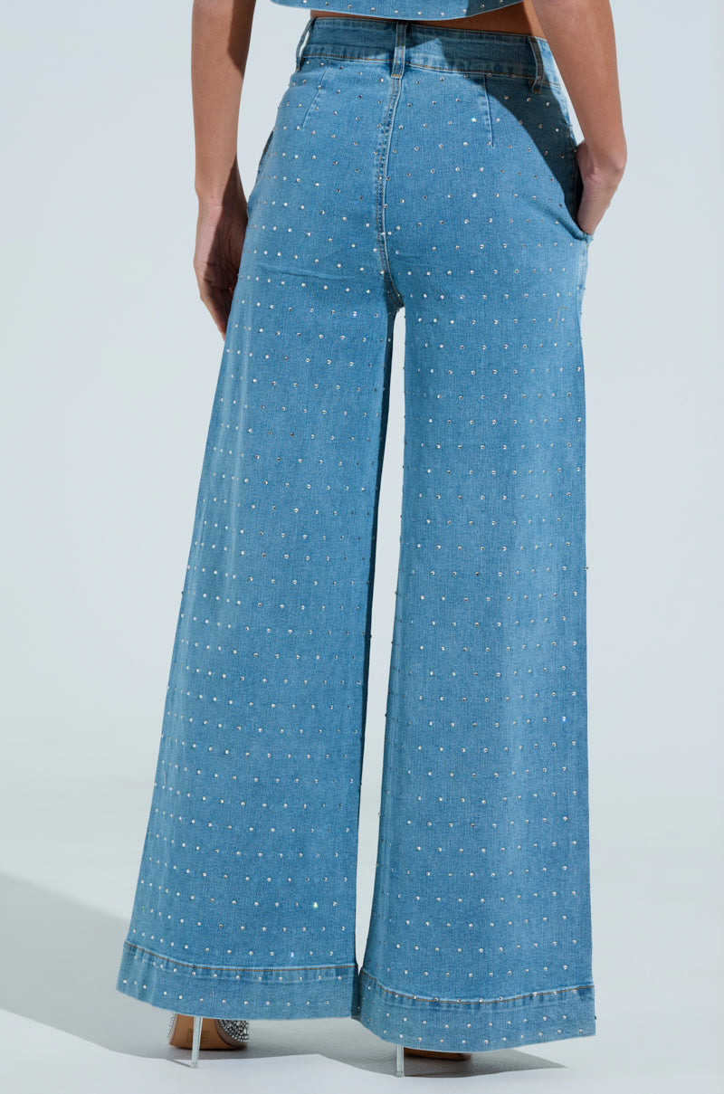 Rear view of someone wearing IT'S GIVING DENIM PANT, featuring high-waisted, wide-leg denim with small silver rhinestones. Paired with light-colored shoes, the look stands out against a plain, light background.