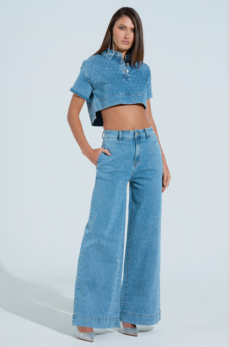 A person poses confidently in light blue denim wide-leg jeans adorned with rhinestones, matching a crop top. Sparkling high heels add flair, while the plain light background enhances the outfit's focus. Product: IT'S GIVING DENIM PANT.