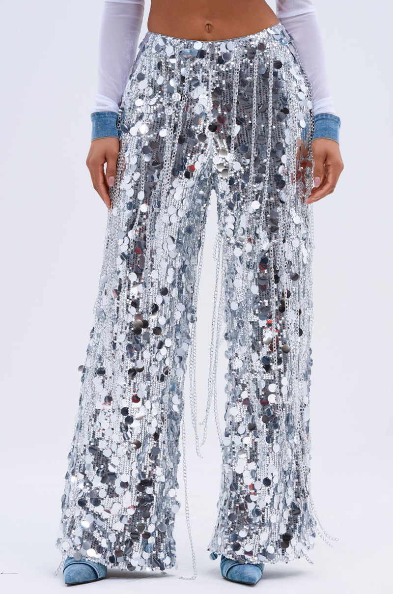 Clad in EXPENSIVE TASTE STRETCHY SEQUIN CHAIN PANTS that glimmer at every step, someone exudes confidence. The outfit, enhanced by a sheer white long-sleeve top and denim shoes against a plain white backdrop, effortlessly captures bold style and glamour.
