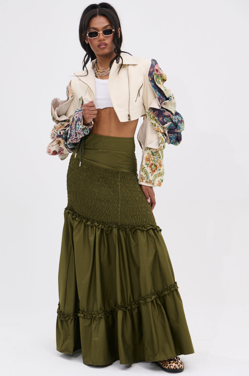 A woman poses confidently wearing a cropped cream jacket with colorful sleeves, white crop top, sunglasses, leopard print shoes, and the IT ONLY GETS BETTER RUCHED MAXI SKIRT in olive green for chic boho vibes.
