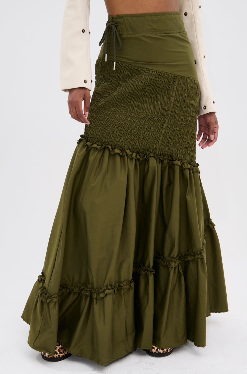 Wearing the IT ONLY GETS BETTER RUCHED MAXI SKIRT in olive green with ruffled tiers and a smocked top, paired with a cream long-sleeve, this look channels boho vibes against a simple background.