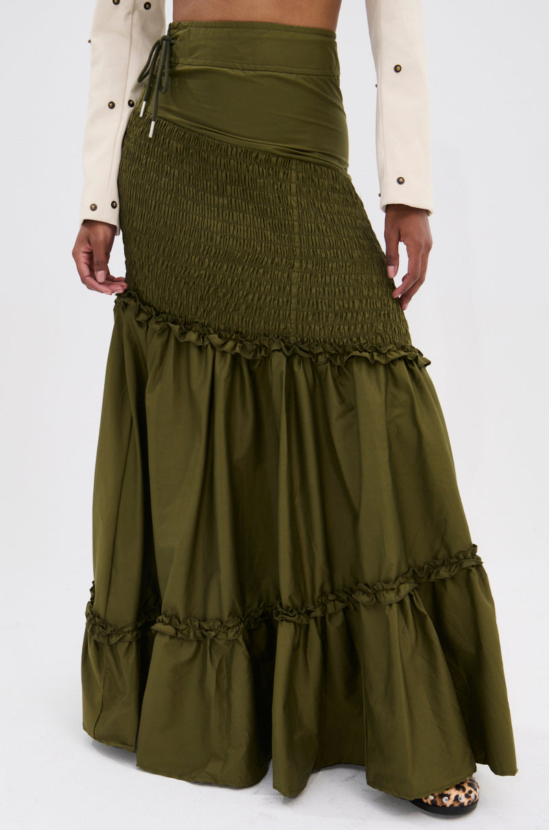 Only the lower torso and legs are visible, showcasing the IT ONLY GETS BETTER RUCHED MAXI SKIRT in olive green with an asymmetrical high-waist and tiered ruffles, paired with a white long-sleeve crop top featuring metal accents.