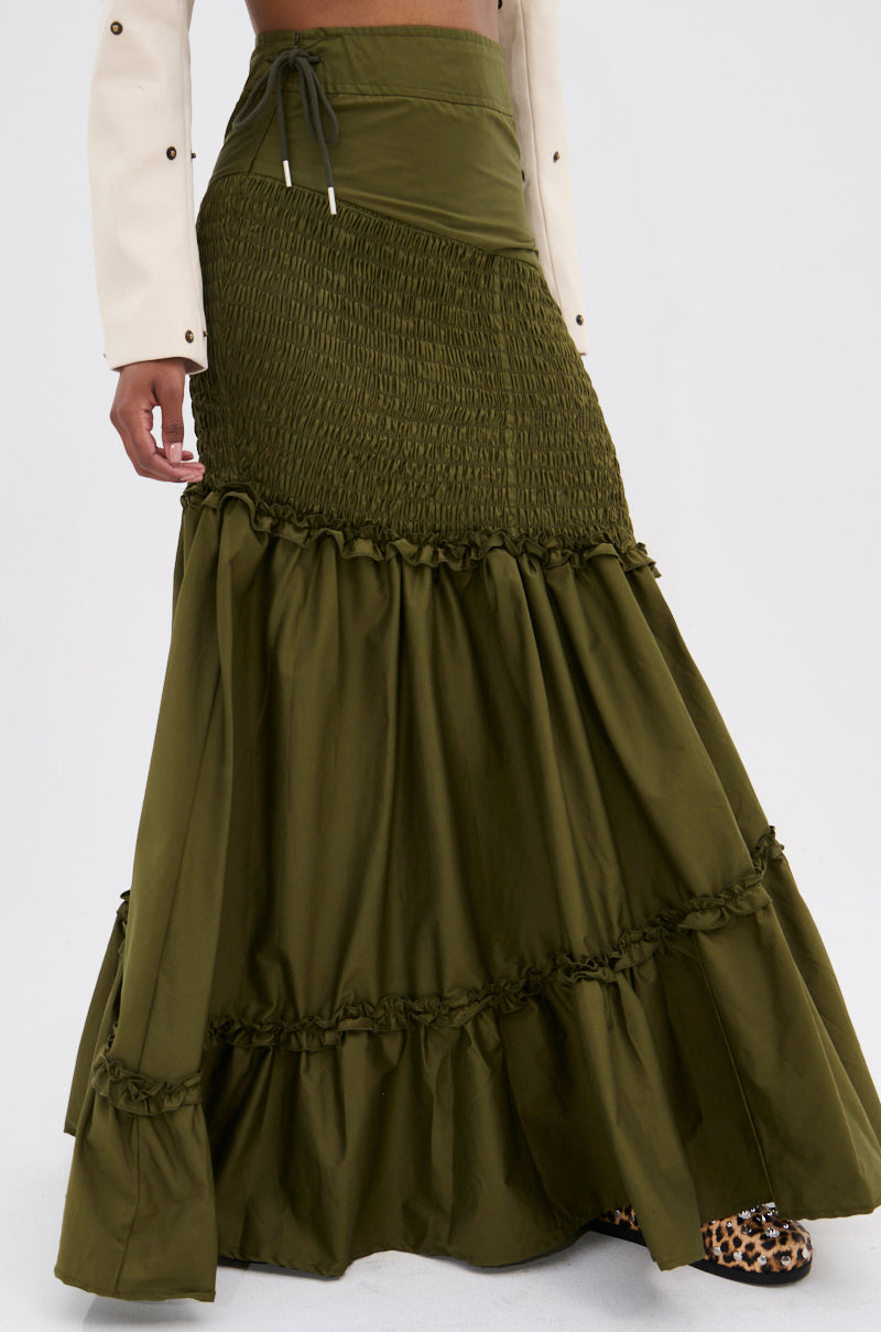 A person wears the IT ONLY GETS BETTER RUCHED MAXI SKIRT in olive green with an asymmetrical high-waist and ruffle details, paired with a white long-sleeve top and leopard-print shoes, showcasing chic boho vibes from the waist down.