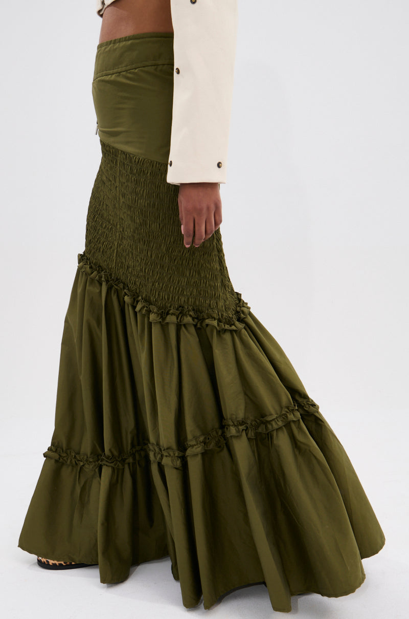 A person stands sideways wearing the IT ONLY GETS BETTER RUCHED MAXI SKIRT in olive green, featuring a high waist, ruched tiers, and boho flair. Paired with a white jacket, only their lower torso and legs are shown.