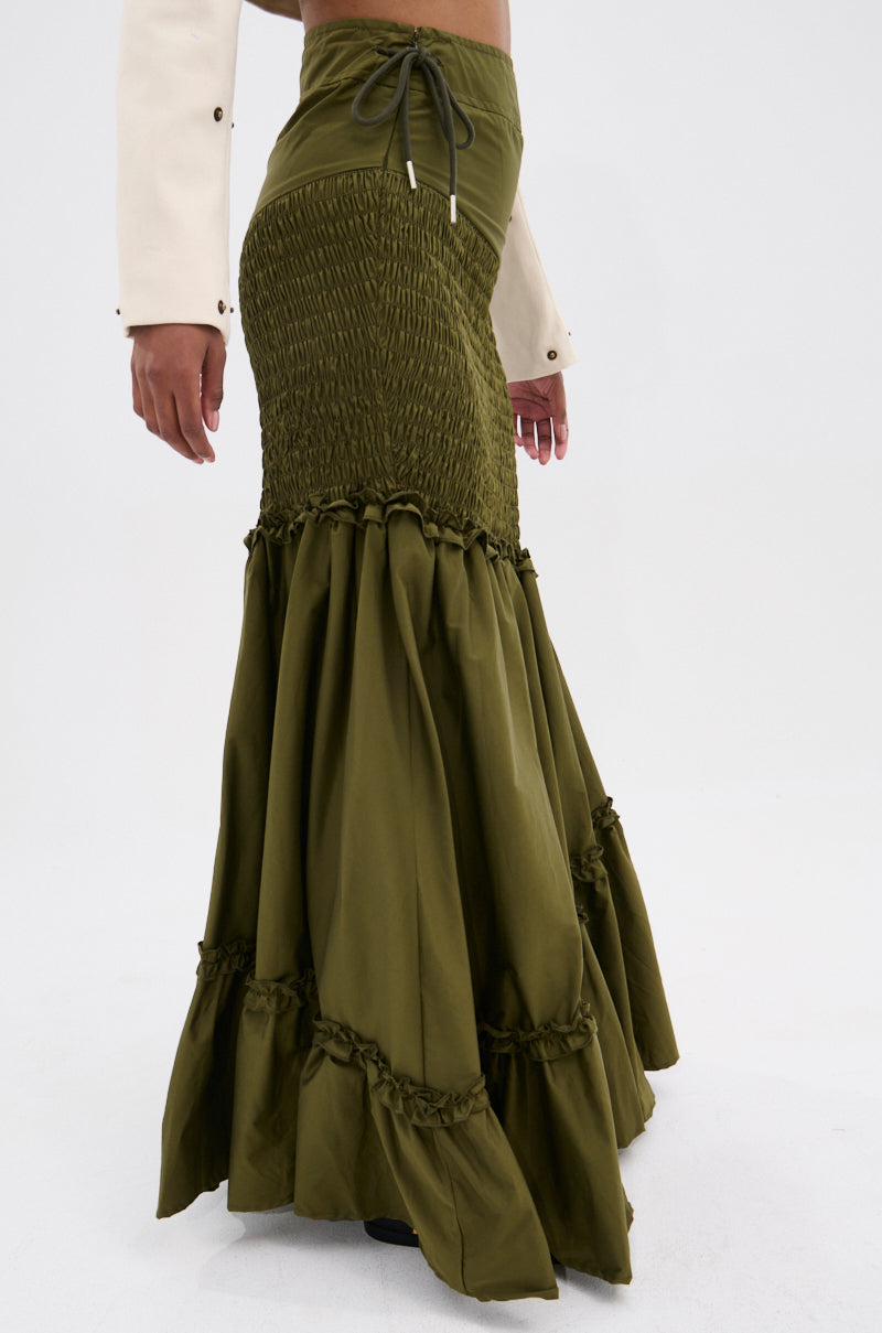Wearing the IT ONLY GETS BETTER RUCHED MAXI SKIRT in olive green with a flared, ruffled hem and cream long-sleeve top, this outfit exudes boho vibes. The high-waisted skirt pops against the plain white background.