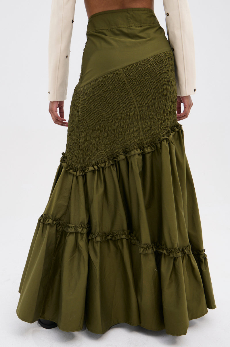 A person is seen from behind wearing the IT ONLY GETS BETTER RUCHED MAXI SKIRT in olive green, featuring a tiered, asymmetrical high-waist boho design, paired with a beige long-sleeve top against a plain background.