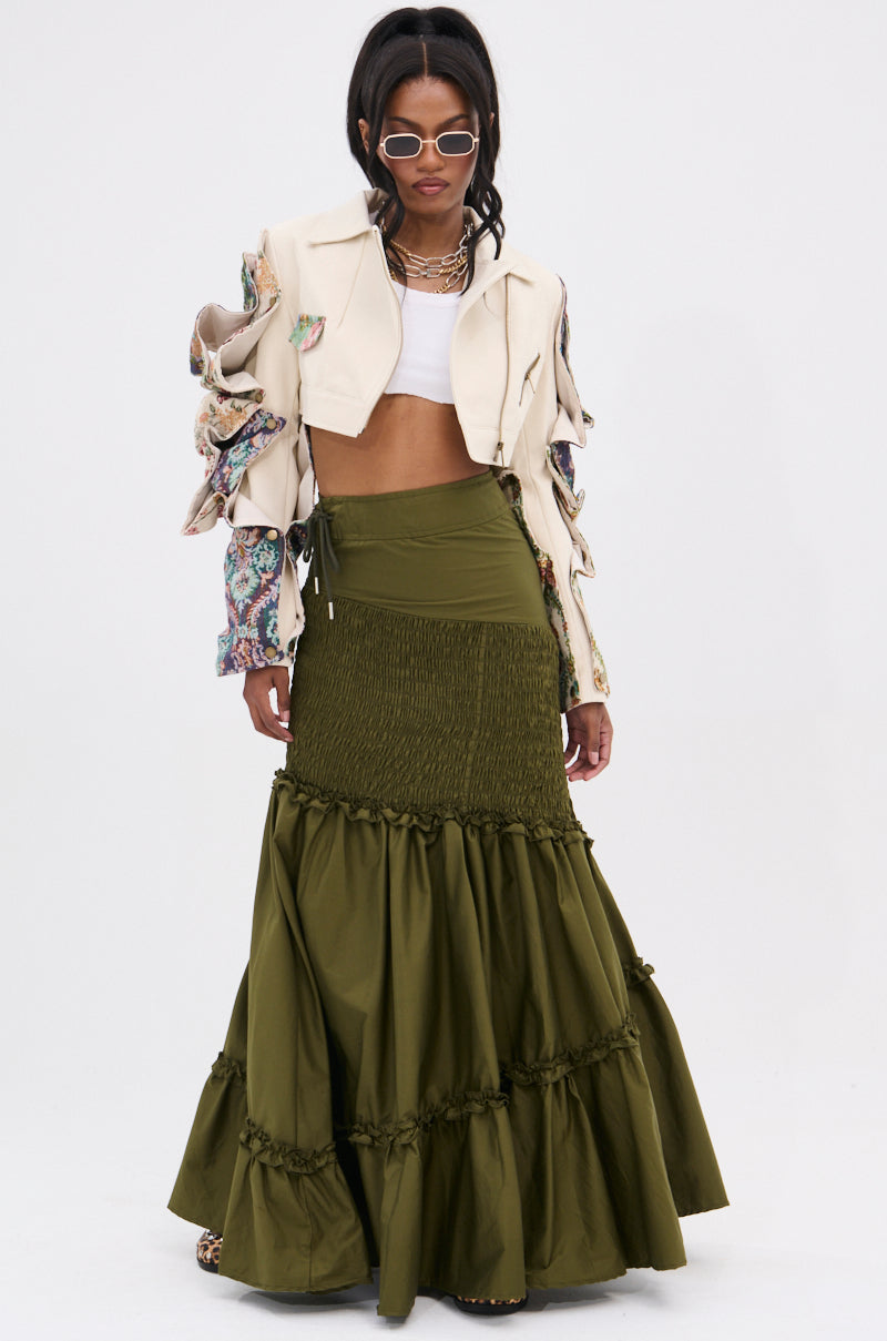 A woman in white sunglasses, a cream jacket with patterned sleeves, a white crop top, and the IT ONLY GETS BETTER RUCHED MAXI SKIRT in olive green poses against a plain white background, radiating effortless boho style.