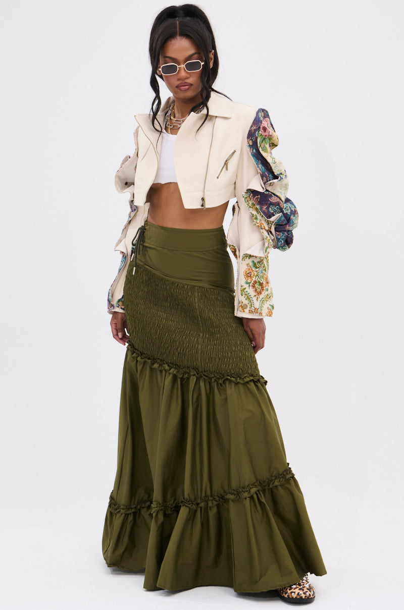 A woman poses in a white crop top, cream cropped jacket with colorful floral sleeves, and the IT ONLY GETS BETTER RUCHED MAXI SKIRT in olive green. She accessorizes with sunglasses, a necklace, and leopard print shoes against a plain white background.