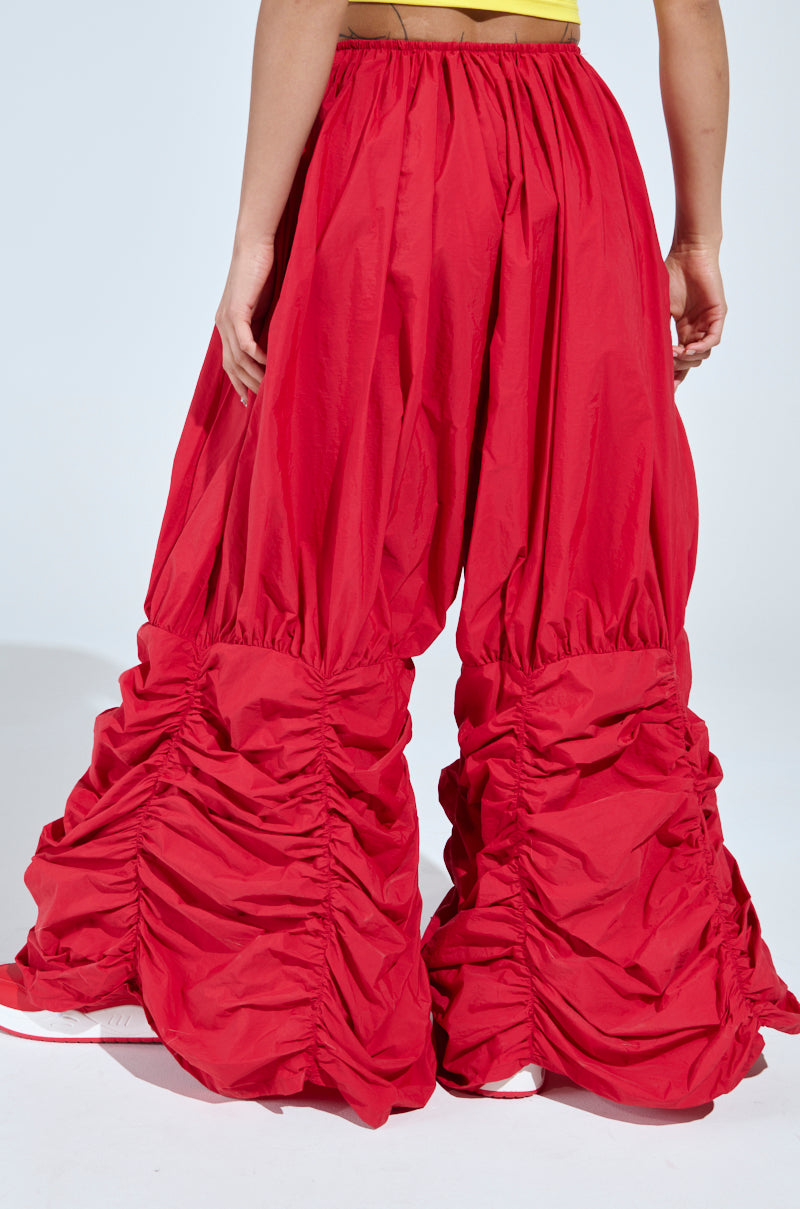 A person wears the WIDE LEG SCRUNCHED HEM PANTS in bright red, featuring a high-rise fit, ruched legs with scrunched hems, and wide flared bottoms, paired with white sneakers. The image is cropped from the waist down.