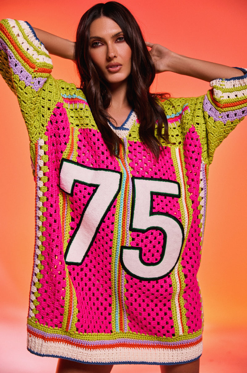 A woman poses with her hands behind her head, wearing the COURTSIDE OR SEASIDE CROCHET JERSEY DRESS, an oversized colorful piece featuring the number 75. The background displays a gradient of orange and pink hues.