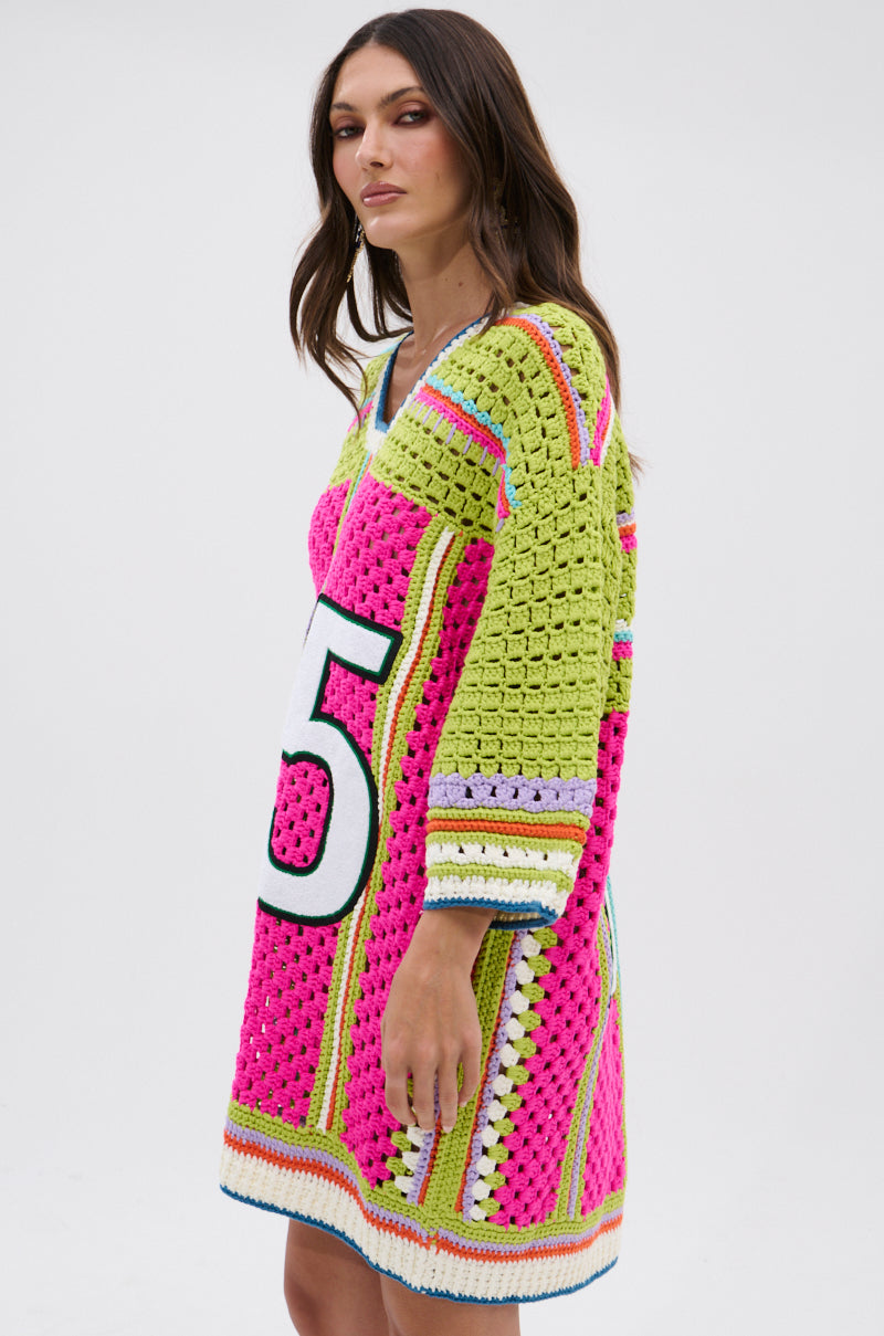 A woman with long brown hair and a neutral expression stands sideways, wearing the COURTSIDE OR SEASIDE CROCHET JERSEY DRESS—a colorful, loose-fitting crochet piece featuring pink, green, yellow, blue patterns and a bold black and white number "5" on the front.