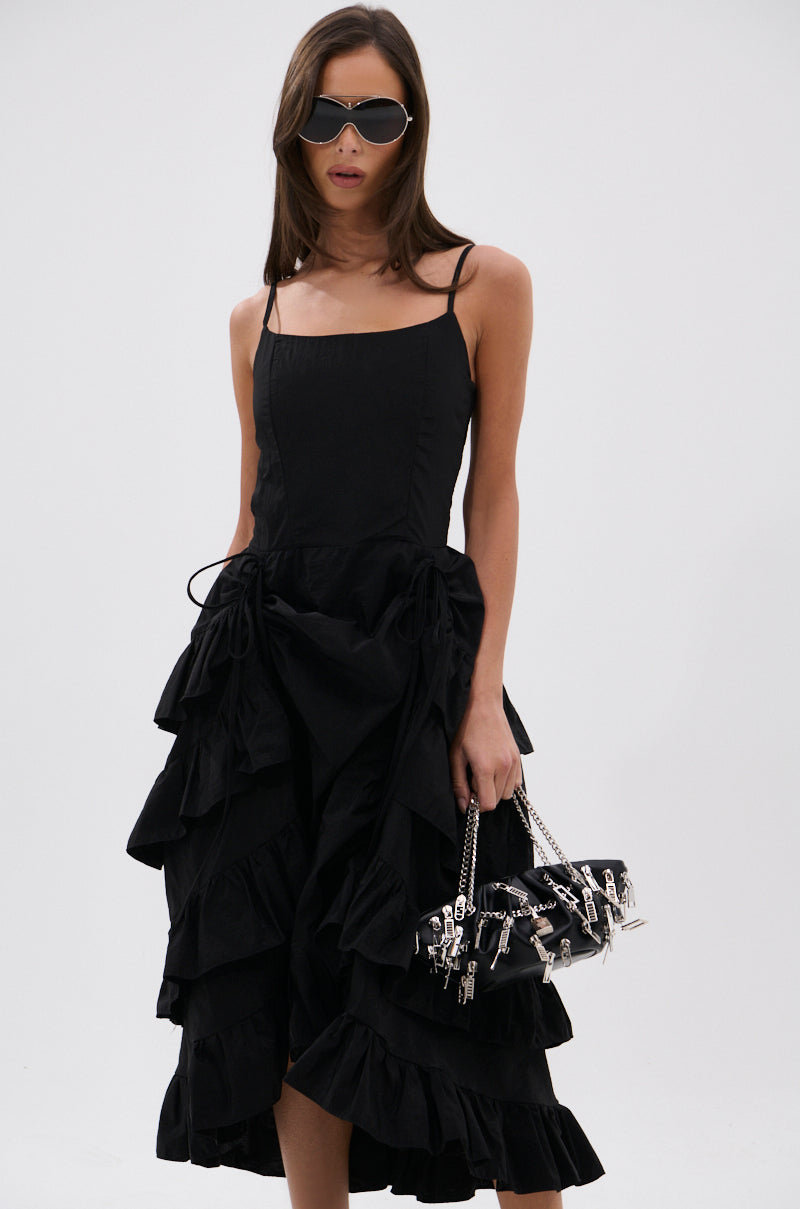 A woman in the ADDED DRAMA RUFFLED MIDI DRESS IN BLACK, paired with dark sunglasses and a black handbag featuring silver chain and charm details, stands against a plain light background.