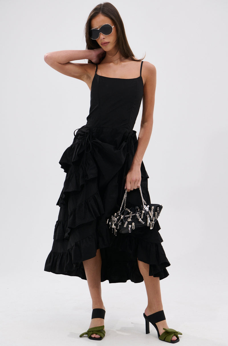 A woman poses confidently in the ADDED DRAMA RUFFLED MIDI DRESS IN BLACK, featuring adjustable spaghetti straps. She pairs it with sunglasses, a black metallic-detailed handbag, and green open-toe heeled sandals against a white background.