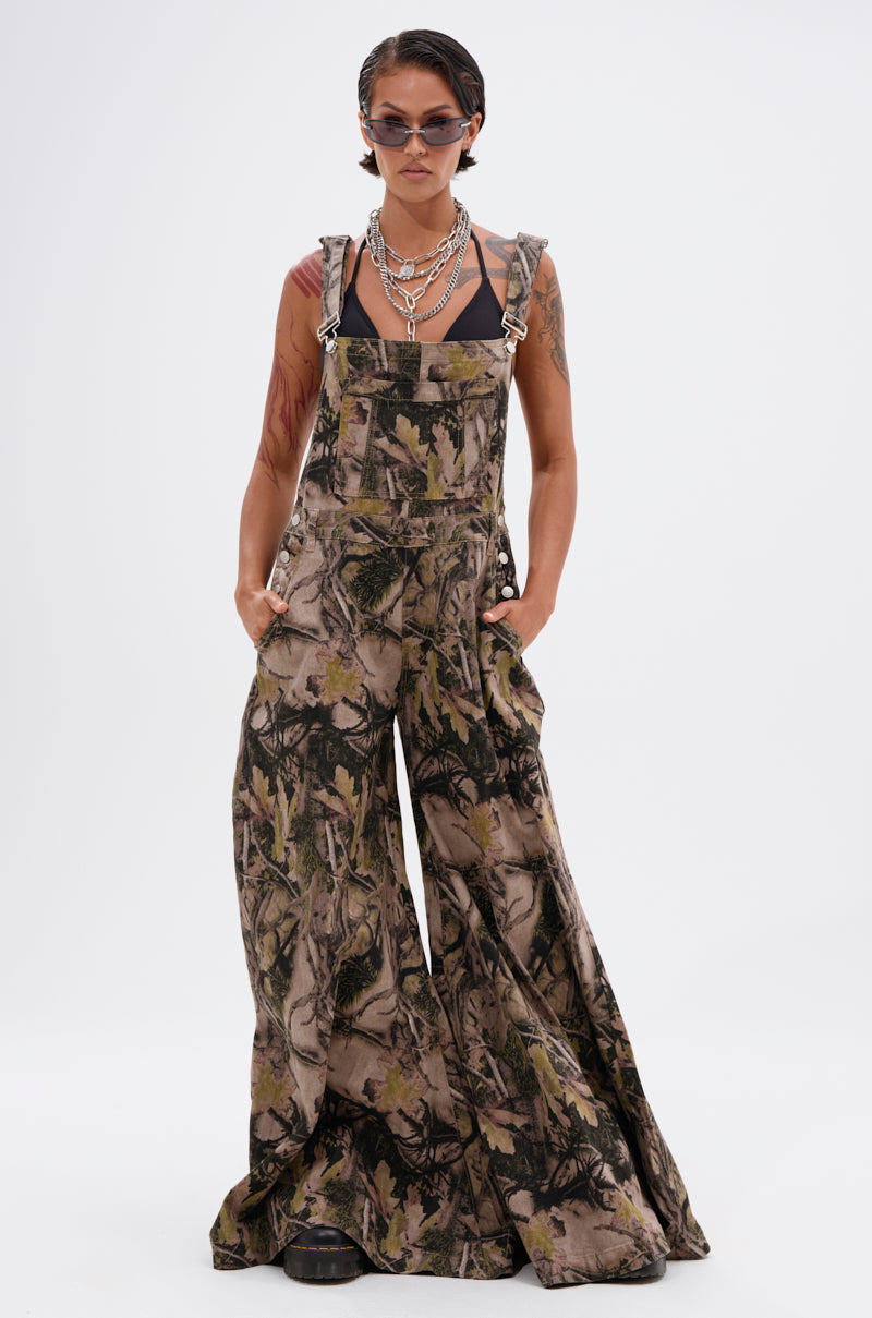 A model in CARMINE CAMO OVERSIZED OVERALLS, a black sleeveless top, chunky boots, layered necklaces, and sunglasses poses with hands in pockets against a plain white background.