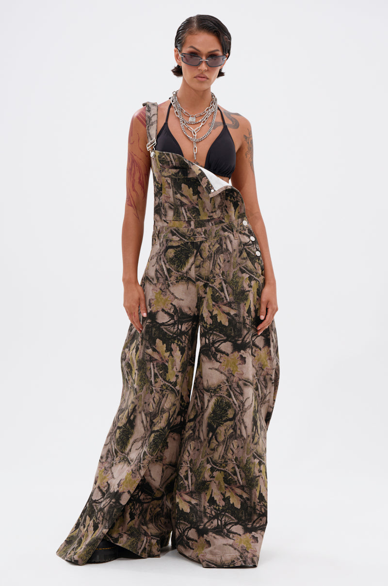 A person in CARMINE CAMO OVERSIZED OVERALLS, statement sunglasses, layered silver necklaces, and a black bikini top stands against a white background.