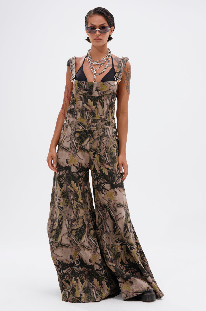 Wearing CARMINE CAMO OVERSIZED OVERALLS over a black top, styled with layered necklaces, sunglasses, and black platform shoes, a person poses against a plain white background.