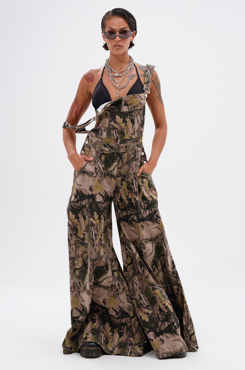 A person models the CARMINE CAMO OVERSIZED OVERALLS with one strap undone, paired with a black bikini top, layered necklaces, sunglasses, and dark lipstick, posing hands-in-pockets against a plain white background.