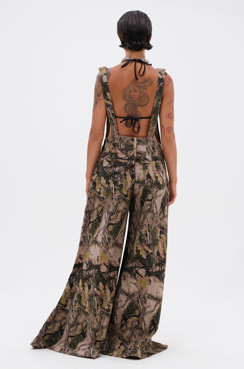 A person with short dark hair and visible tattoos models the CARMINE CAMO OVERSIZED OVERALLS, featuring an open back and forest camo print, paired with a black halter top against a plain white background.